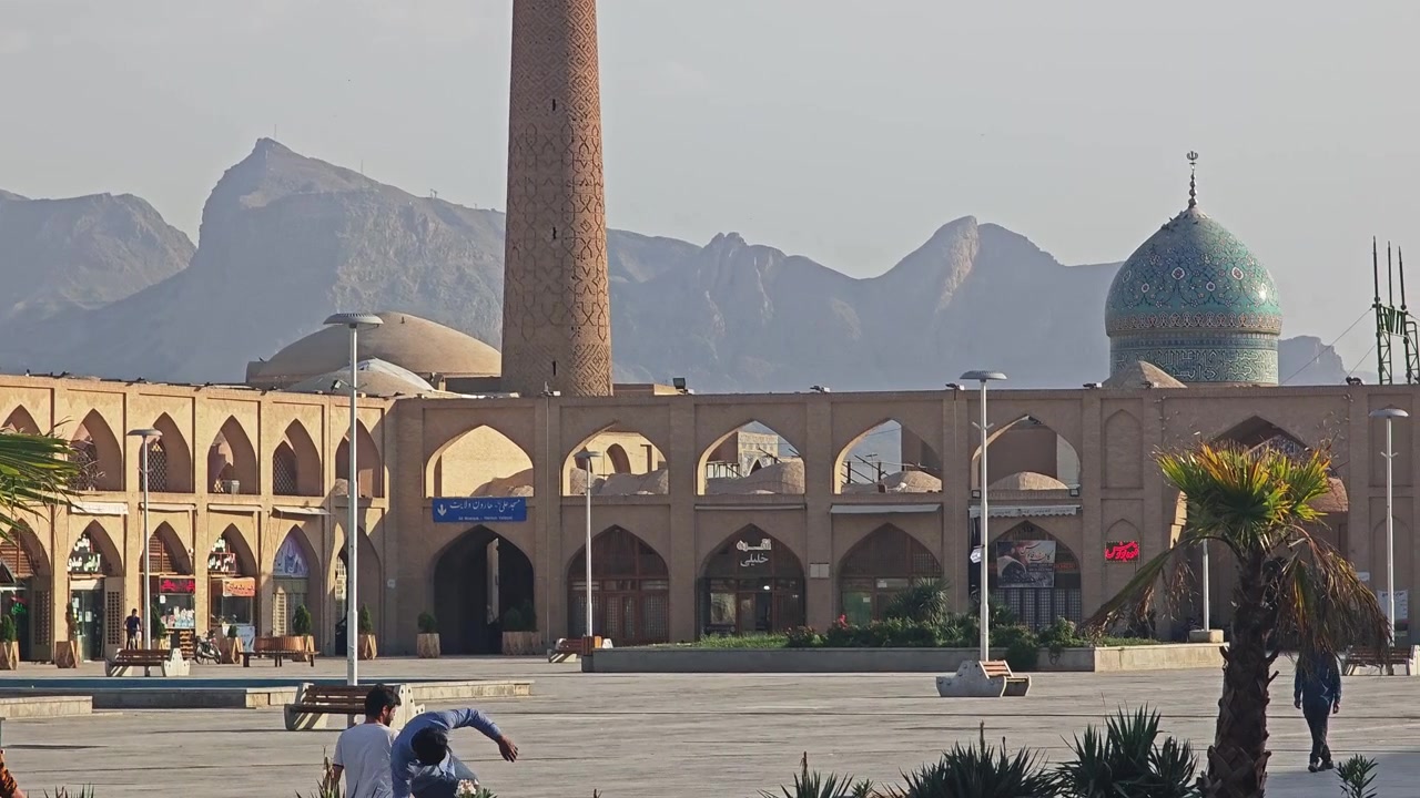 Pessoas caminhando em frente a uma mesquita histórica e montanhas no Irã.