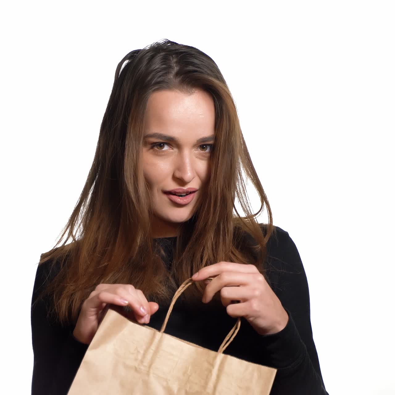 A beautiful woman in a black sweater looks mysteriously in a paper packbag and throws it in frustration and go away on the white background. Purchases. Shopping