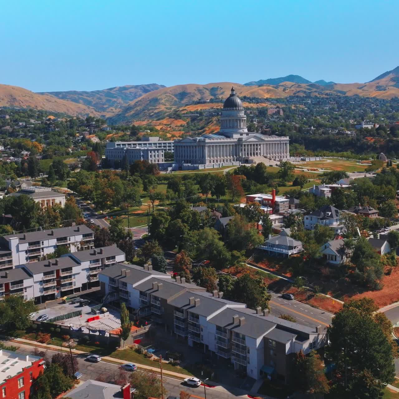 Beautiful Utah State Capitol surrounded by greenery and modern houses. Brown bare mountains and clear blue skies at backdrop