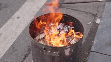 Orange-flame-in-metal-bucket,-Vietnamese-offering-ritual-of-burning-votive-paper,-slow-motion