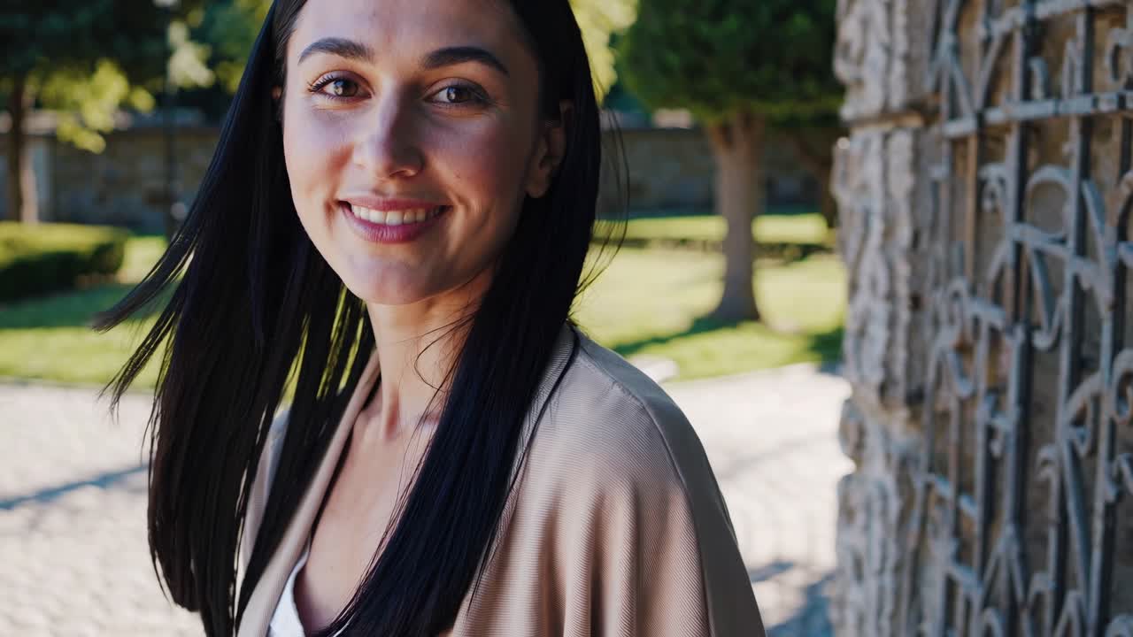 Confident young woman standing near ornamental gate, wearing light brown jacket, exuding relaxed charm in sunlit urban park setting with soft, natural background