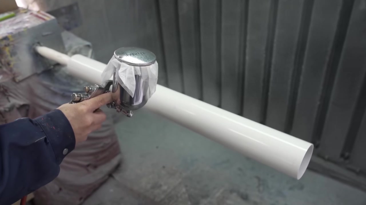Arbeider spraymaler en hvit teleskoprørkropp med en håndholdt sprøytepistol inne i en industriell lakkboks.