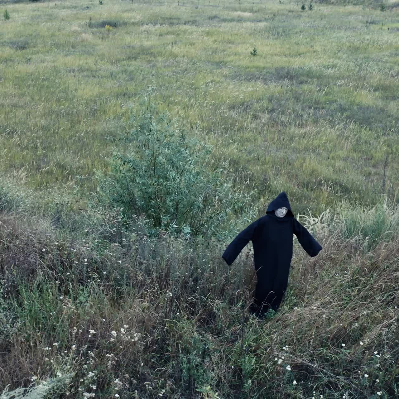Top view of a dark spooky figure in grass. Ghost in black mantle with hood standing alone in the field. Halloween time.