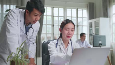 hospital medical doctor's office: two asian professional physicians talking, consulting and working on laptop computer. specialists discuss patient online treatment, writing digital medicine prescriptions