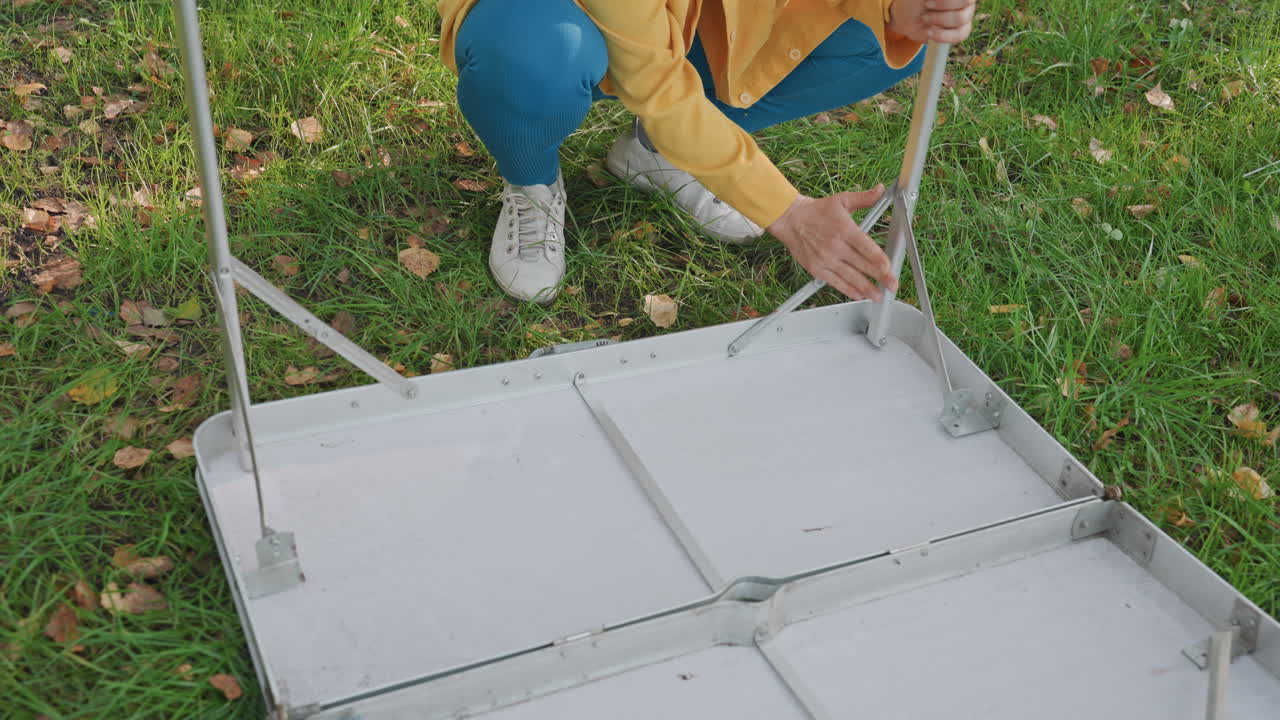 Folding camping table on grass, solo camper kneels and unfolds aluminum table, secures legs and hinges, overcast sky, green lawn, casual jacket and sneakers, hands adjusting latches, compact gear