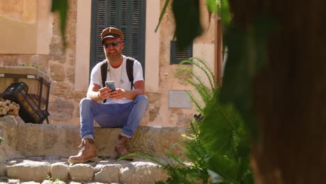 male traveler sitting near aged stone house on mobile phone