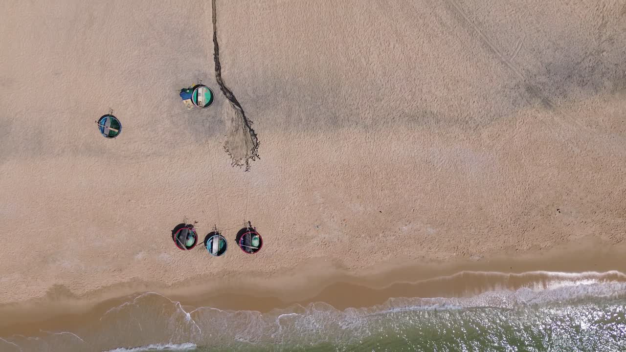 Drone flies sideways with a top-down view in Mui Ne, Vietnam. Fisherman buckets are parked on the beach, shoreline centered, with waves gently hitting the sand.
