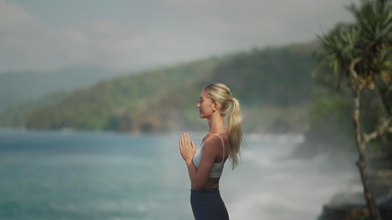 Fit blond yoga female model standing in relaxing zen meditation with powerful crashing waves at Bali coast, slowmo 120fps