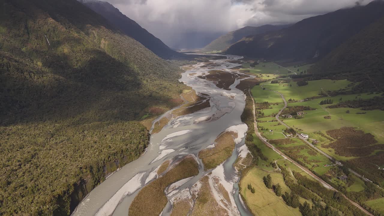 Drone luftfoto følger Bealey-floden, der løber gennem en bred alpedal nær Arthur's Pass på New Zealands Sydø, med grønne marker, bjerge og dramatiske skyer