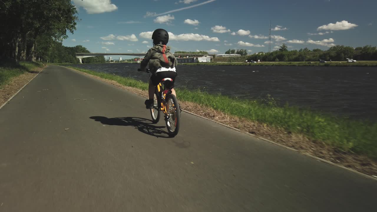 Little boy rides bike on paved path along lake, POV follow, 4k
