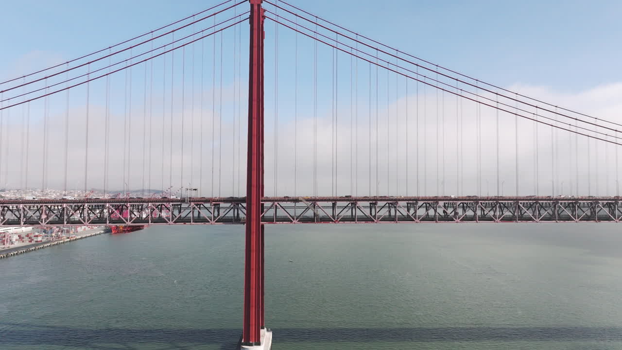 Aerial drone shot towards the 25th April Bridge in Lisbon, Portugal, Europe. Sunny and bright with clouds in the background. Cars driving on bridge, side view. Drone flying backwards