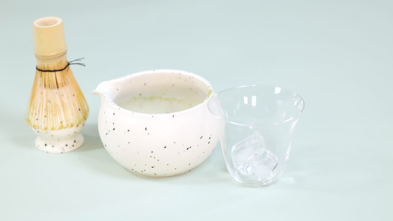 Hand places ice cubes in glass beside matcha bowl and bamboo whisk, bright studio lighting