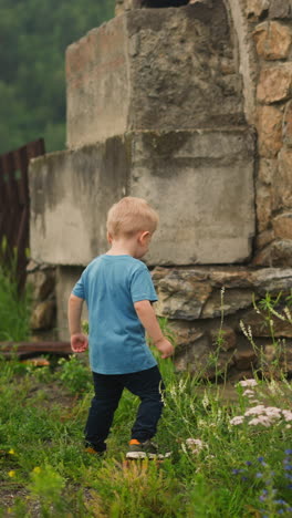 clumsy little boy in blue t-shirt lifts up on wooden ground for braizer in green field in summerhouse yard on spring day backside view slow motion