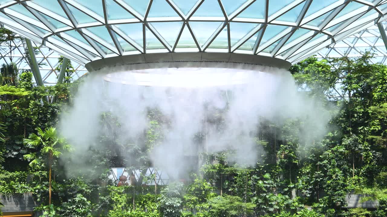 Dense mist pours from a circular indoor waterfall structure, surrounded by tropical plants under a geometric glass dome, with bright natural daylight and stationary camera