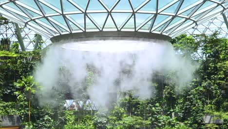 dense mist pours from a circular indoor waterfall structure, surrounded by tropical plants under a geometric glass dome, with bright natural daylight and stationary camera
