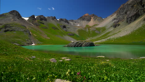 aerial cinematográfica vista amplia cuenca del lago de hielo isla de silverton lago agua azul agua clara tundra alpina impresionante cordillera de montañas flores silvestres mediados del verano de día bluesy hermoso movimiento lento hacia la izquierda