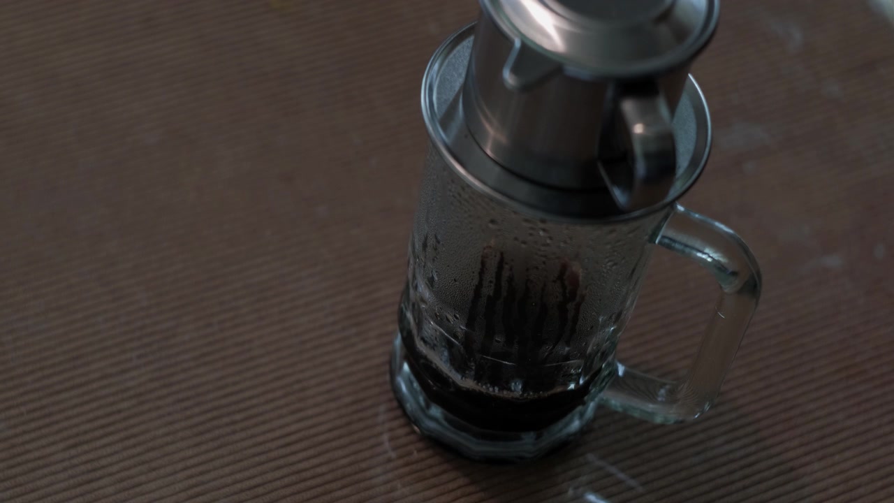 Static shot of a Vietnamese phin filter brewing and dripping dark coffee into a glass mug on a table indoors