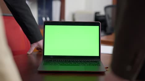primer plano de portátil con pantalla verde en la mesa con gente de negocios irreconocible alrededor. dispositivo chromakey en la oficina en el interior con hombres y mujeres analizando en línea. tecnologías modernas y negocios electrónicos.