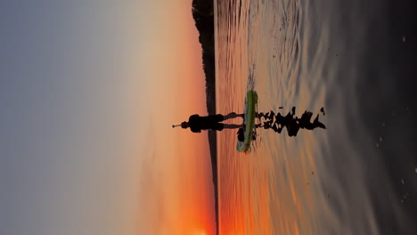 rear view of a man moving on the sup board by the river. beautiful orange setting sun over the water at backdrop. vertical video.