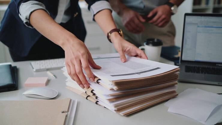 Reaching for pages, woman in dark blazer flipping and stacking manila folders at desk for review