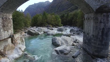 Fliegen-Durch-Doppelbogen-Steinbrücke-Im-Verzasca-Tal-In-Der-Schweiz-Luftaufnahme-Des-Kristallklaren-Türkisfarbenen-Bergflusses