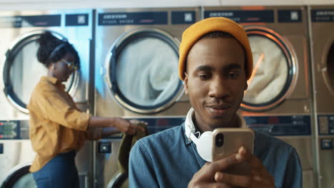 close up of stylish man in yellow hat tapping and texting message on smarphone while sitting in laundry service room