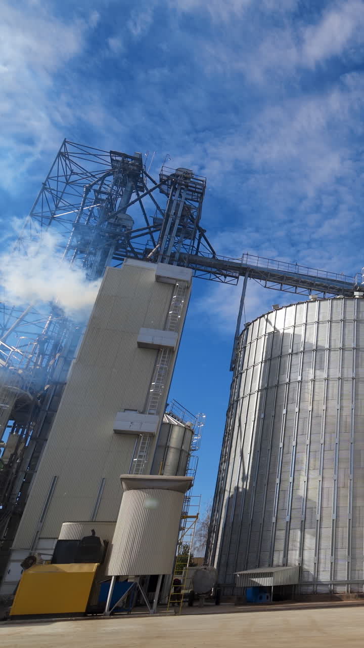 Harmful smoke filling the atmosphere from industry. Aluminum grain elevators on the modern agricultural factory at bright sunny day. Vertical video