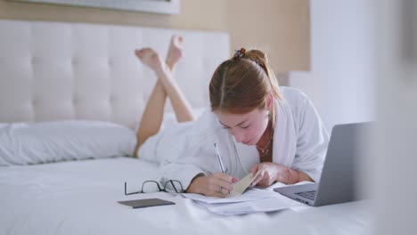 Young-lady-doing-homework-assignment-lying-on-bed-with-netbook