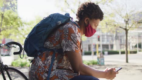 African-american-man-in-city,-wearing-face-mask-sitting-using-smartphone-in-street