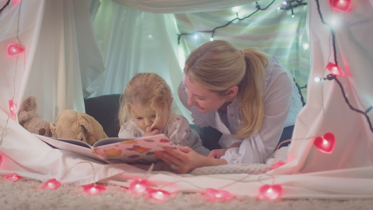 Mother and young daughter reading story in homemade camp in child's bedroom at home - shot in slow motion