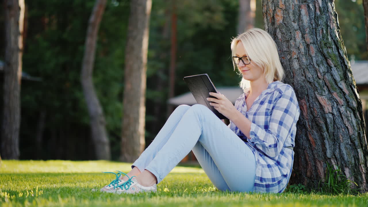 A Young Woman Is Using A Smartphone Sits On The Grass Under A Tree In The Backyard Of The House 4k V