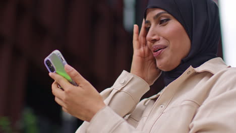 close up of muslim businesswoman wearing hijab standing outside modern office getting good news about job offer or promotion on mobile phone