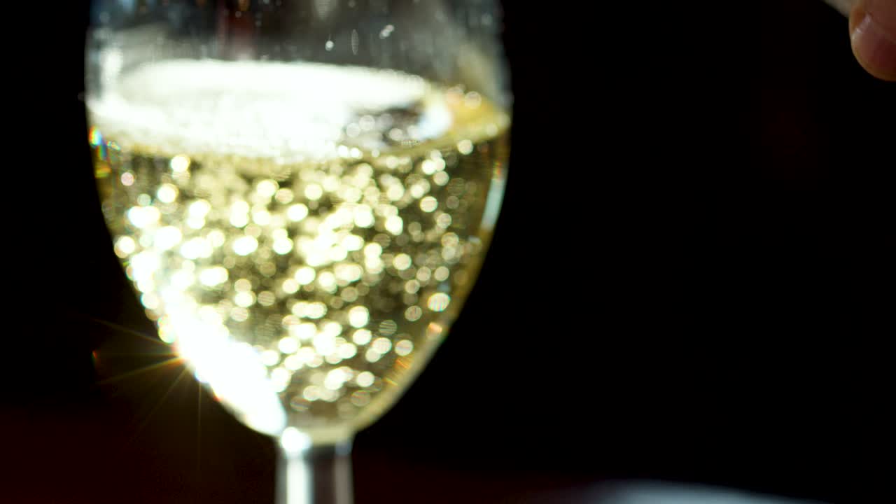 Sparkling white wine poured into clear glass, golden bubbles rising, strong sunlight, dark background