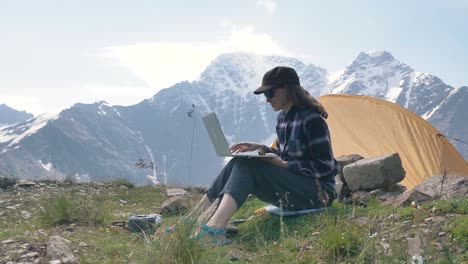 a freelance woman works at a laptop outdoors against the backdrop of high snowy mountains next to a tent