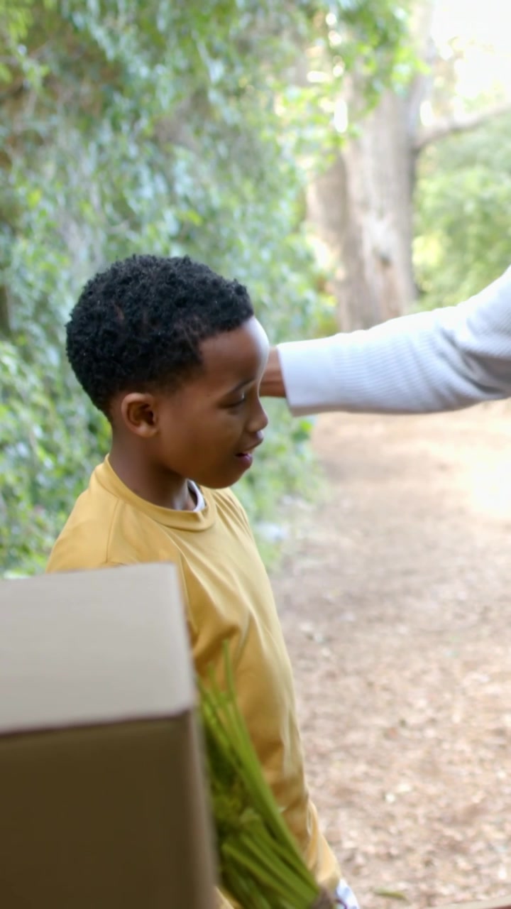 Video verticale: Bambino con maglietta color sabbia che batte il cinque alla mano di un adulto che mostra un grembiule, offrendo assistenza vicino a una cassetta di verdure su un sentiero.