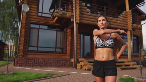 a slender and beautiful woman in sports clothes with an open press is preparing to start training on the lawn near her home stretching her arms and shoulders with warming up exercises