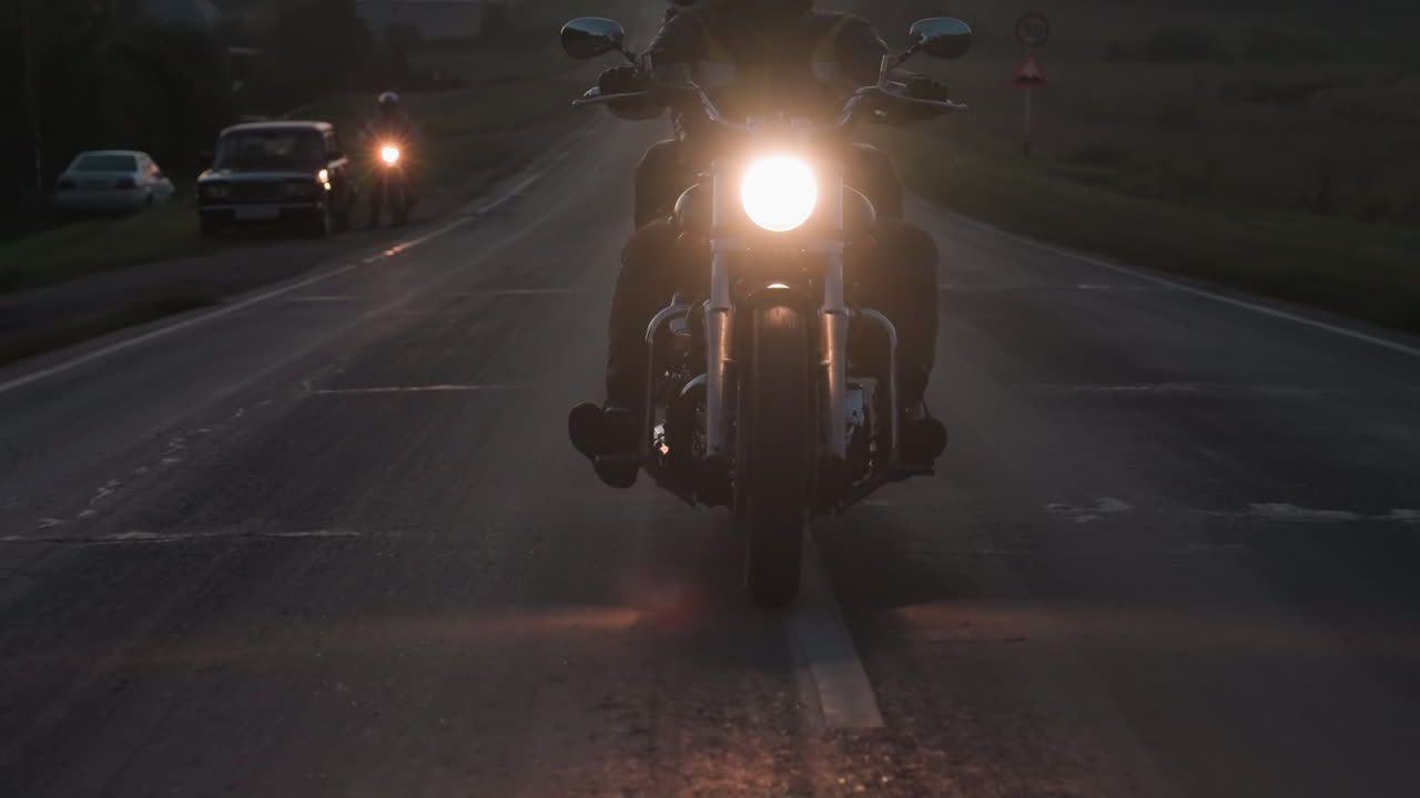 Motociclista che percorre una strada di campagna durante una pattuglia serale con un faro luminoso che illumina la superficie screpolata e l'orizzonte luminoso mentre il motociclista supera i veicoli parcheggiati