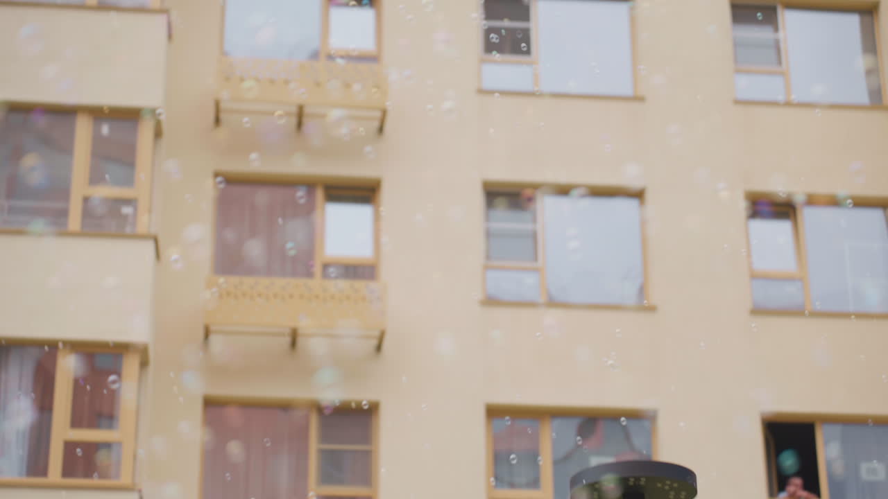 Floating soap bubbles drift across courtyard near brick building and windows, gentle motion around lamppost with fence and grass, calm outdoor scene with subtle reflections