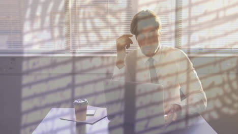 Animation-of-plants-and-rain-on-window-over-tired-caucasian-businessman-using-laptop-at-office-desk