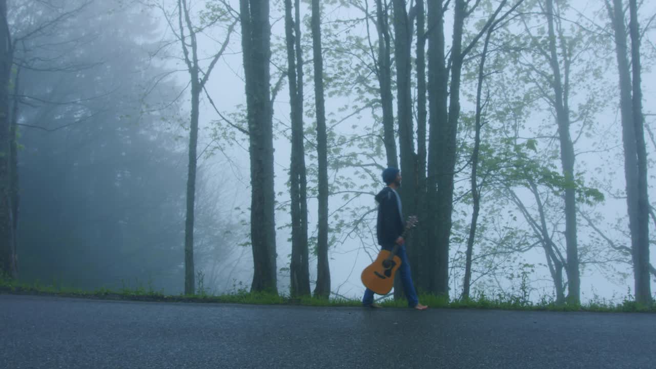 Una persona cammina sul ciglio della strada con la sua chitarra in una foresta nebbiosa