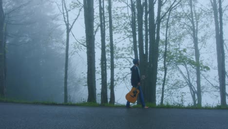 Una-Persona-Camina-Al-Lado-De-La-Carretera-Con-Su-Guitarra-En-Un-Bosque-Nublado