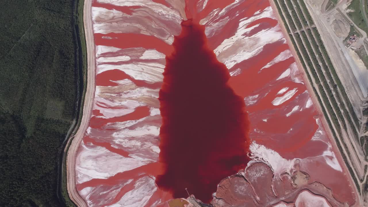 Luftfoto af en enorm rød mudderdam ved kysten, et industrielt affaldsprodukt fra aluminiumoxidproduktion. Billedet fremhæver reservoirets enorme omfang med stærke kontraster mellem r