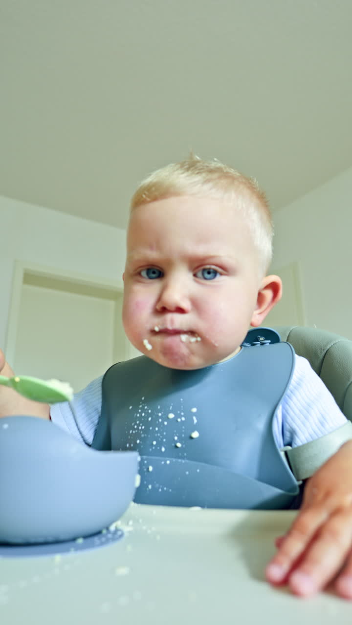 Blond 2 year old boy eats milk porridge with a spoon. Toddler sits at home at a table and eats porridge from a silicone plate. Children's tableware