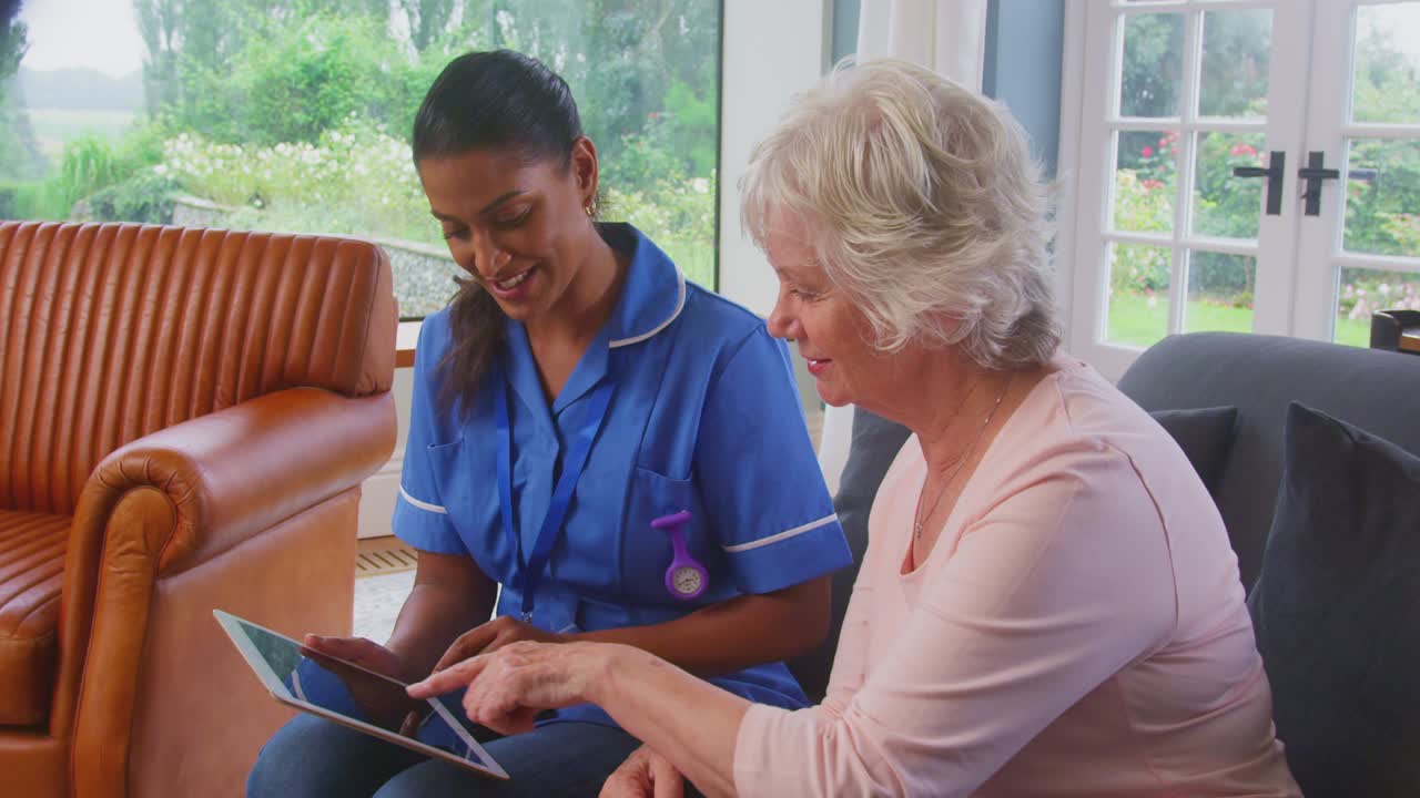 Senior Woman At Home Talking To Female Nurse Or Care Worker In Uniform Using Digital Tablet