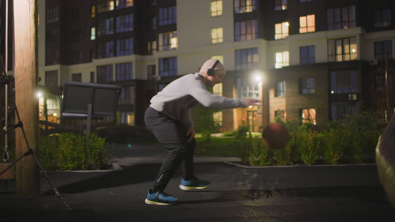 Uomo che si allena a basket di notte sotto le luci di un appartamento, palleggiando con sicurezza sul pavimento bagnato del cortile, con felpa, scarpe da ginnastica e cuffie, espressione concentrata, allenamento urbano solitario