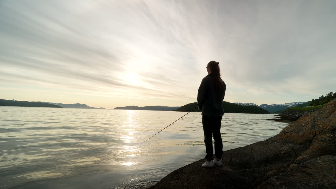 Female on Tromso coastline casts into fjord as she is spin fishing at sunset