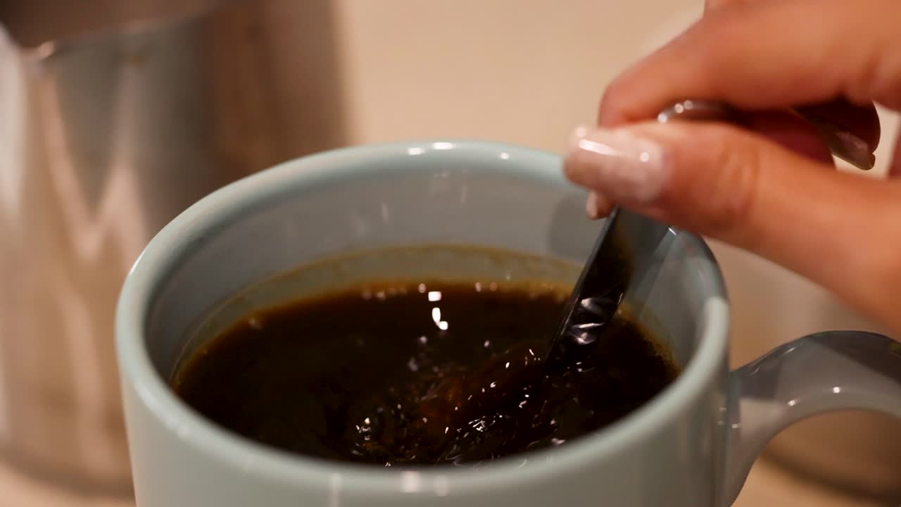 A hand stirs coffee in a blue mug, creating swirling patterns. Warm lighting enhances the cozy kitchen atmosphere