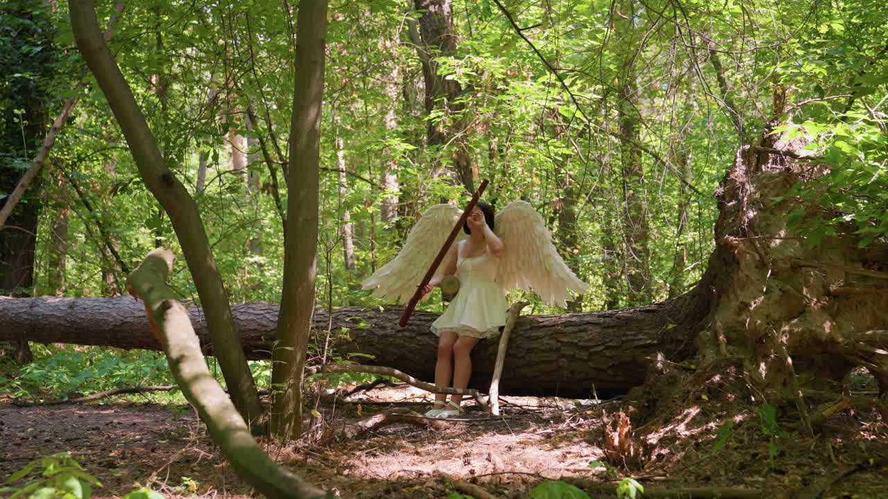 Mystical angel woman with white wings sits on fallen tree holding long wooden staff surrounded by lush forest greenery, sunlight filtering through leaves