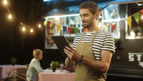 Young-Man-Waiter-In-Apron-Smiling-And-Using-Tablet-Device-In-Evening-At-Food-Truck-In-Festive-Park