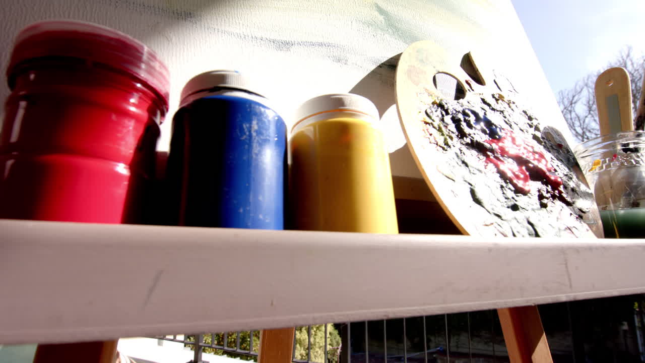 Shelf with colorful paint cans and palette faces a canvas